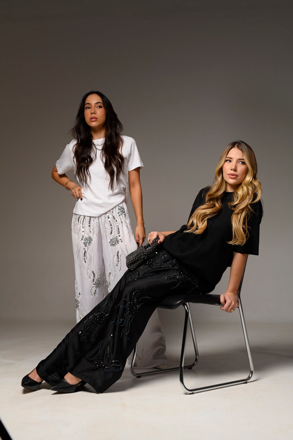 Duas modelos com calças bordadas à mão da Viis, uma sentada em cadeira de estúdio